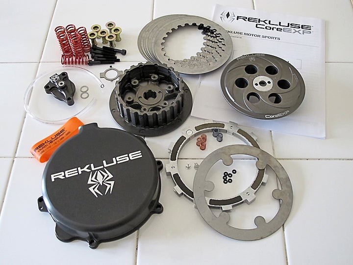 Rekluse’s Core EXP auto-clutch kit includes (clockwise from upper left) new pressure plate springs/bolts/retainers, new drive plates, instruction manual, new pressure plate, EXP disk with optional springs, new (slightly deeper) cover, rubber band for tuning free play, and new adjustable master cylinder (for hydraulic clutches – cable clutches get an adjustable pressure plate).  At center is a beefy new billet aluminum hub.
